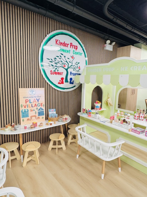 Play village area featuring prominent Kinder Prep Enrichment Center signage with round table and wooden stools